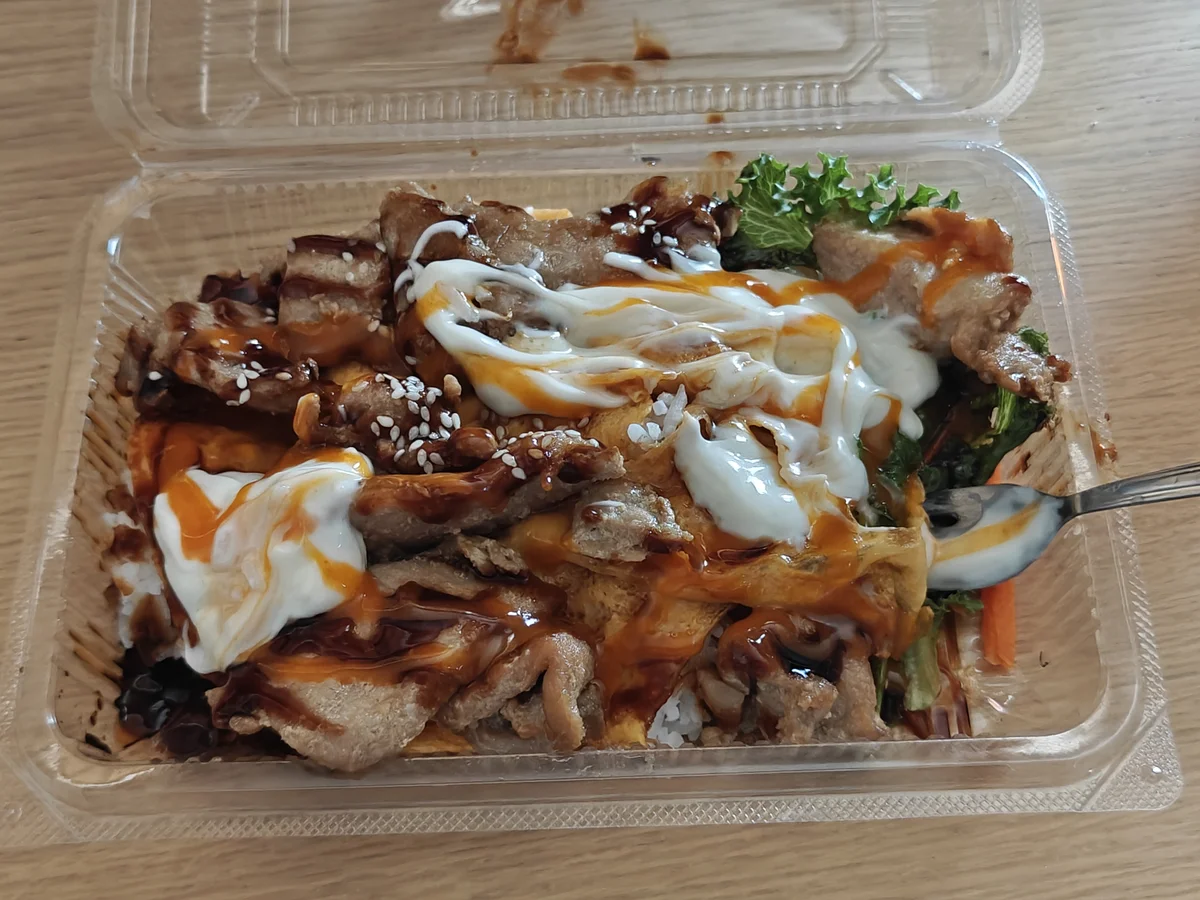 A plastic clamshell container doing its best to contain what appears to be a rice bowl having an identity crisis — teriyaki, sriracha mayo, and regular mayo all showing up uninvited to the same party.